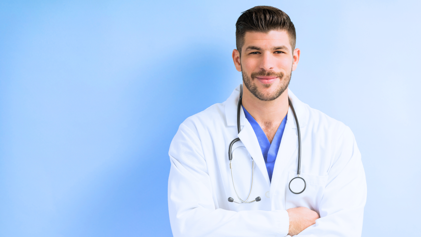 A male doctor stands with arms crossed, wearing a white coat and a stethoscope around his neck, looking confidently at the camera.