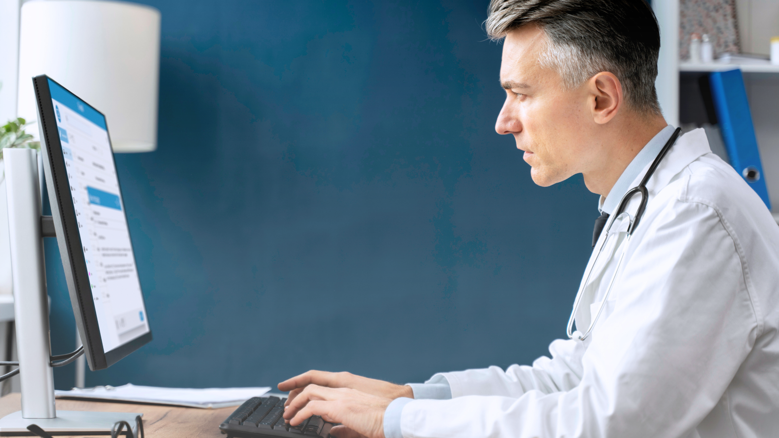 A male doctor in a white coat sits at a desk, working on a computer in a professional office setting.