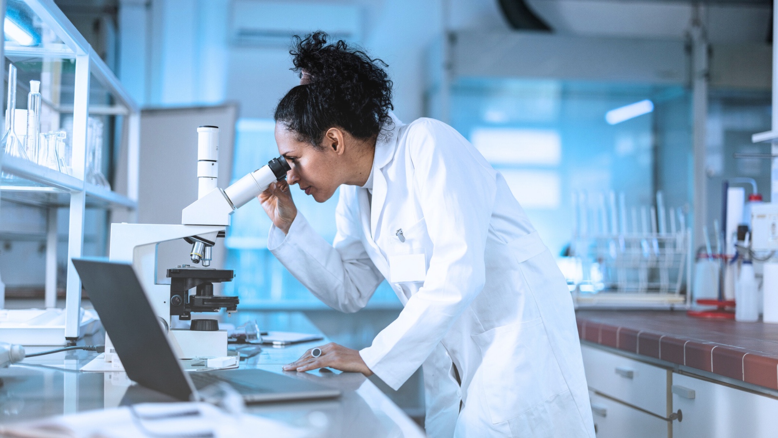 A woman in a lab coat examines a sample through a microscope in a laboratory setting. Next to here is a laptop.