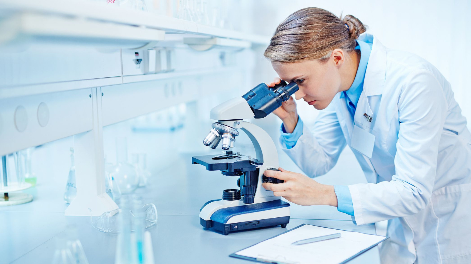 A woman in a lab coat examines a sample through a microscope in a laboratory setting.